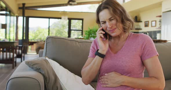 Caucasian pregnant woman in pain sitting on sofa, touching belly and having call