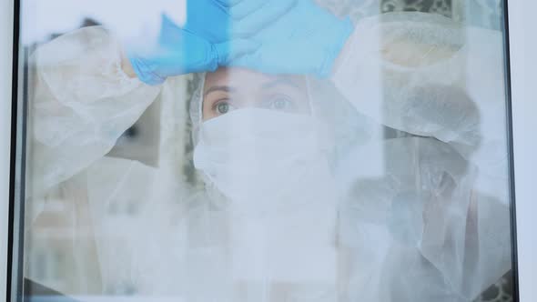Sick woman in white protective suit in medical face mask stares longingly out through window