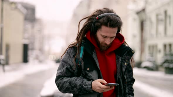 Trendy Dressed Young Man with Dreadlocks is Walking in City and Listening to Music By Wireless