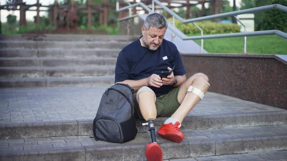 Mature Caucasian Male Amputee with Artificial Legs Sitting on Stairs in City Park Checking Messages