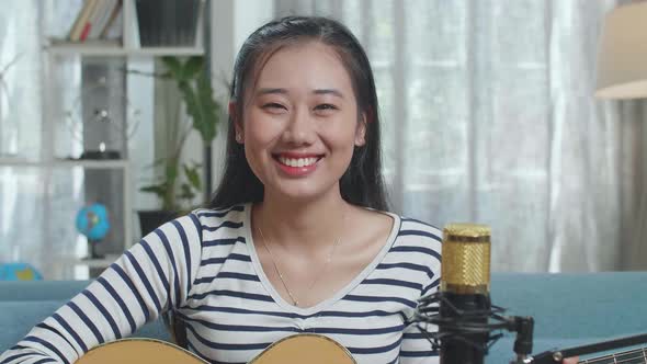 Close Up Of Asian Woman With Microphone Holding A Guitar And Smiling To Camera At Home