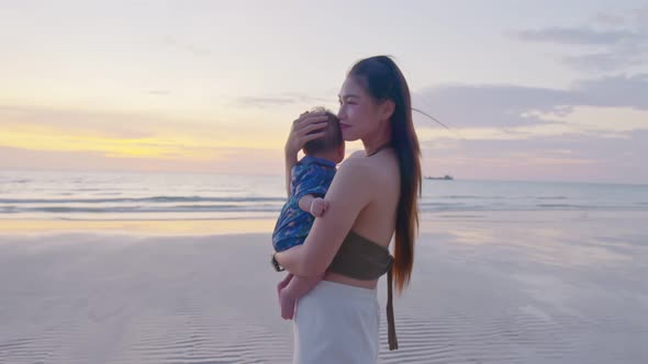 Happy mother with baby boy walks by ocean on the beach in summer