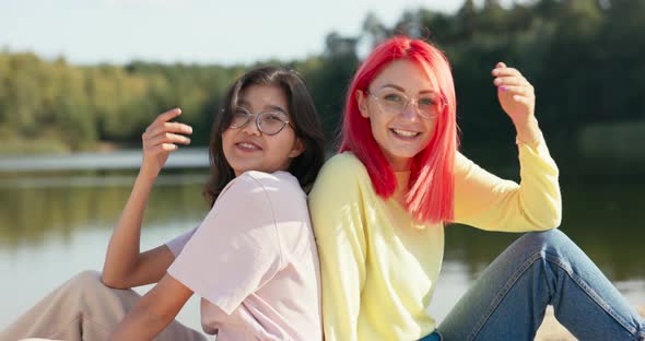 Joyful Smiling Pretty Girls in Glasses are Sitting on the Sand of the Beach Near the Lake Leaning