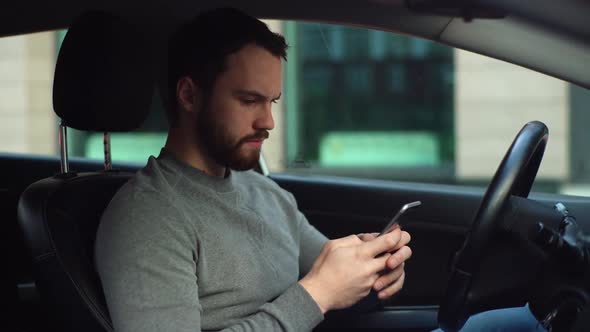 Serious bearded young man sitting in car and typing online message on cell phone, side view.