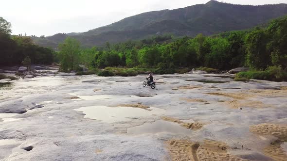 Aerial: Man Driving a Motorcycle on Paved Stone Surface with Sand in the River.