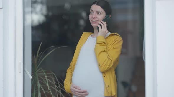 Beautiful Positive Pregnant Woman Talking on Phone Standing at Glass Door Looking Out
