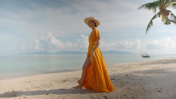 Barefooted Woman in Yellow Long Dress and Hat Taking Solitary Walk By Ocean Touching Hat with Hand