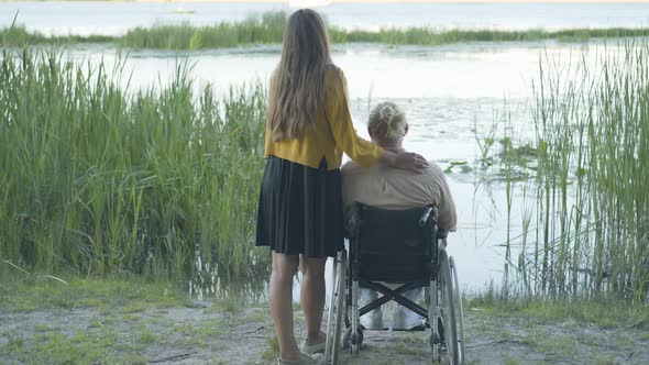 Back View of Caucasian Couple Admiring Sunset on River Bank. Young Woman and Disabled Man on