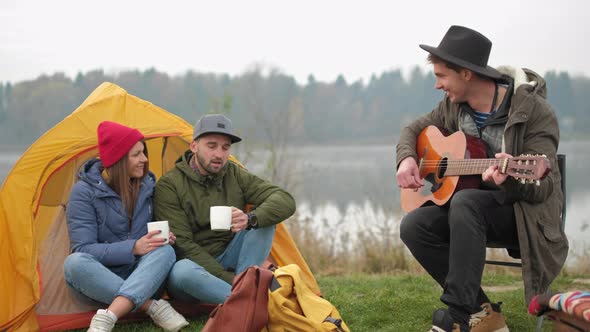 Group of Friends camping,They Are Sitting Around Camp Fire, Playing Guitar