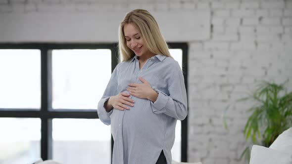 Young Happy Pregnant Woman Caressing Belly Standing in Bedroom at Home and Smiling Looking at Camera