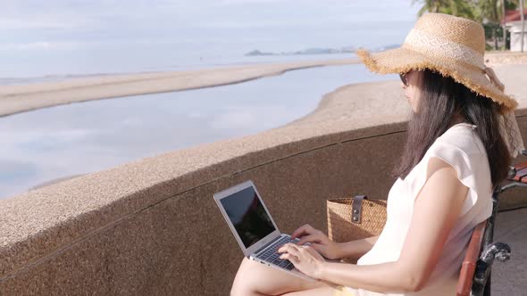 a carefree Asian woman sitting on beach bench by the sea, using laptop computer