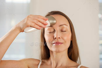 Middle aged woman using anti-aging electric massager doing led therapy facial massage