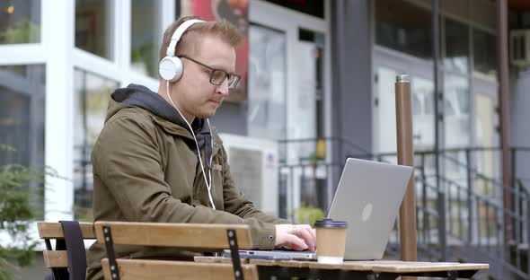 Business man in headphones works online using a laptop while sitting in a cafe on the street. 