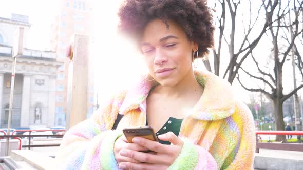 Slow motion young beautiful curly hair black woman outdoor in the city smiling using smartphone