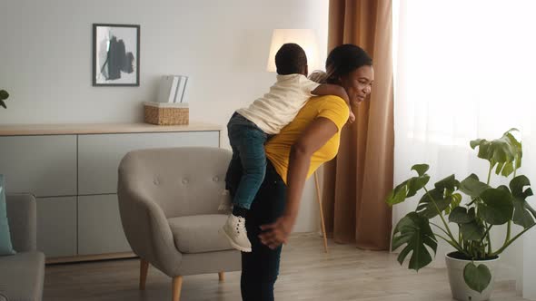 Cheerful Black Mom Making Squats At Home With Little Son On Back