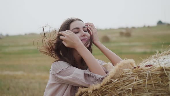 Happy Girl Leaned on Haystack in a Field Correcting Blowing Hair and Smiling