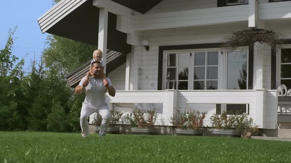 Happy Father Does Morning Exercises Crouches While Holding Son on His Neck