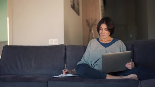 Confident mature woman working on laptop and writing something in notepad