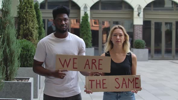 A African American Man and a White Woman Stand on the Street Holding Posters for WE ARE and THE SAME
