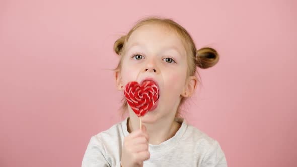 Funny Little Blonde Girl Smiling and Playing with Red Heart Shape Lollipop Caramel on Pink