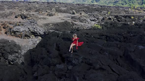 Girl in Red Dress Sitting on Solidified Black Lava Field on Volcano Batur in Bali, Indonesia. Aerial