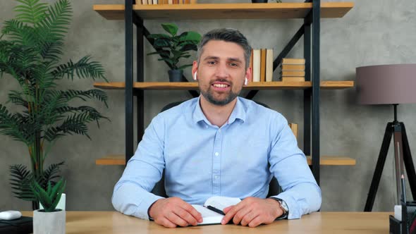 Smiling business man at home office in wireless earphones greets the camera listens nods head