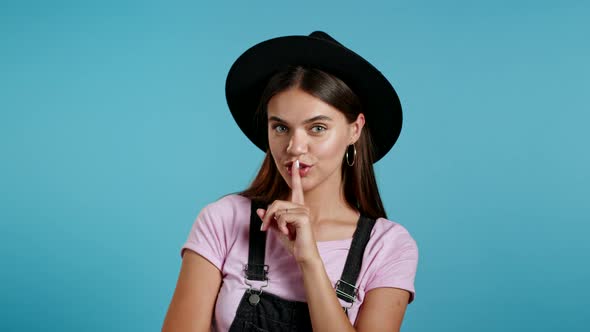 Beautiful Hipster Woman in Hat Holding Finger on Her Lips Over Blue Background. Gesture of Shhh