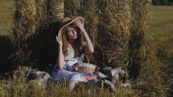 Beautiful Redhead Woman in Dress and Hat Sits Near a Haystack