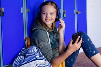 Smiling caucasian schoolgirl with headphones uses smartphone by school lockers