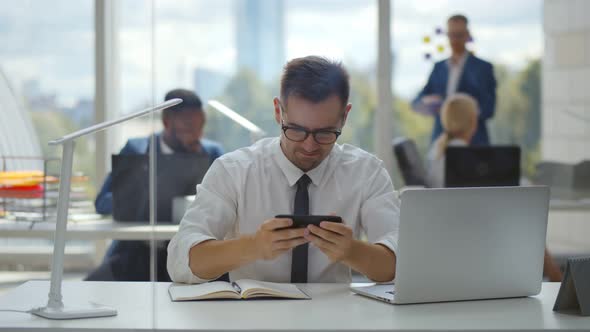 Handsome Young Worker Playing Games on Smartphone Sitting at Desk in Modern Office