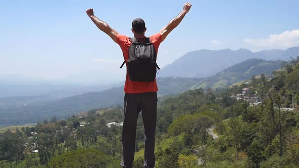 Male Hiker with Backpack Reaches Up Top of Mountain and Outstretched Arms Up. Young Traveler Jogs To