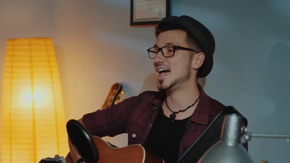 Close Up of Bearded Guy in Hat Emotionally Singing Into Microphone and Playing Guitar in Home Studio