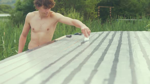 Young man applying sealant into gaps of wooden boat cabin roof planking topless