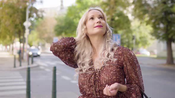 A Middleaged Caucasian Woman Looks Around with a Smile in an Urban Area  a Busy Road