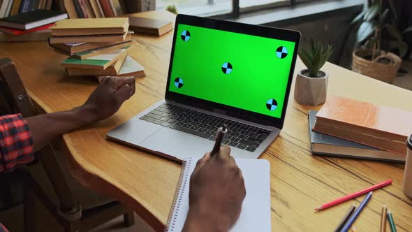 Young Black Man Student Studying Looking on Green Screen of Laptop and Making Notes in Copybook