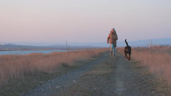 A Girl in a Jacket Runs and Fools Around on the Beach By the Sea with a Rottweiler Dog