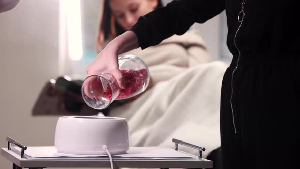 Waxing Master Puts Dry Wax in the Melting Heat Machine While Her Woman Client Reading Magazine