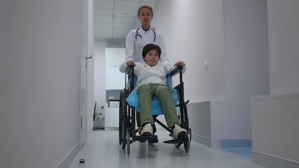 Wide Shot Caucasian Boy in Wheelchair with Serious Doctor Pushing Mobility Aid Device Walking in