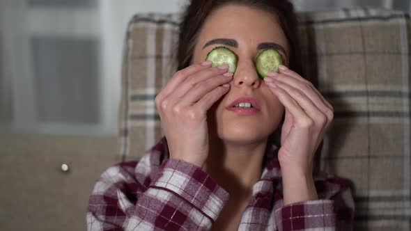 Portrait of Pretty Young Woman in Pajama Making Eye Mask From Cucumbers in Cozy Evening, Chilling