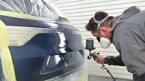 Spraying trunk of blue vehicle by professional painter with protective equipment