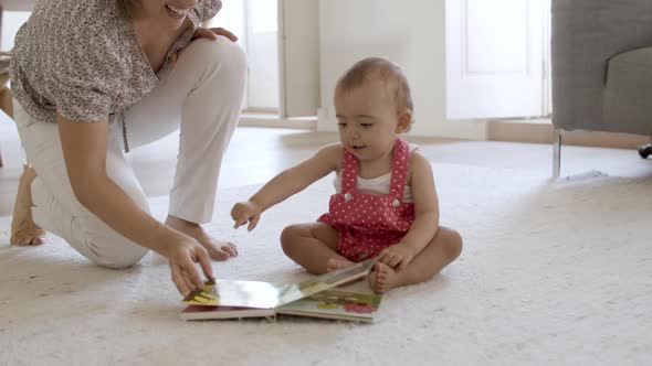 Happy Mom Reading Book with Baby Girl Sitting on Floor