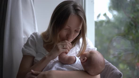 Caucasian Woman Kissing Tiny Hand of Newborn Baby Sitting on Windowsill at Home Indoors Smiling