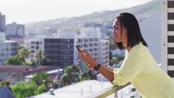 Mixed race gender fluid person drinking cup of coffee and using smartphone on roof terrace