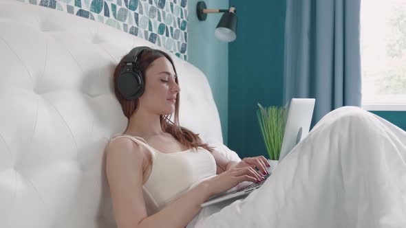 Confident Brunette Woman in Headphone Typing Message in Computer on Bed