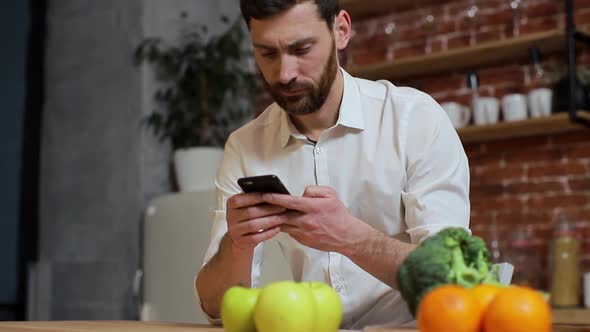 Man Browsing on Mobile Phone at Home Kitchen. Handsome Young Man Browsing on Smartphone Smiling