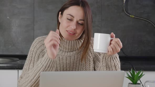 Millennial Young Lady in Woolen Sweater Laughing Is in Correspondence on a Computer at Home Drinking