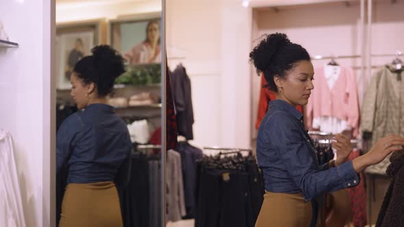 Beautiful African American Woman Choosing Clothes Hanging on Hangers in Trendy Clothing Store