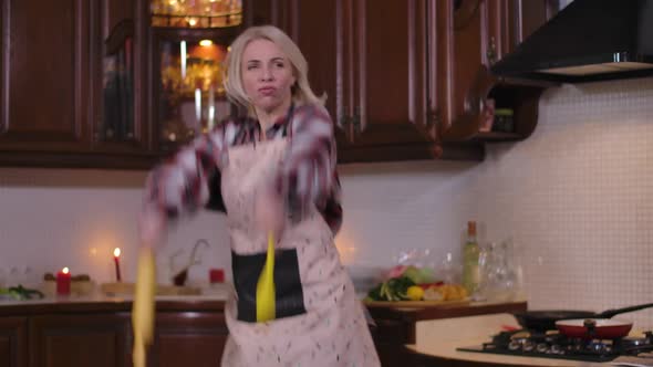 Portrait of Carefree Cheerful Young Woman Dancing with Rolling Pin and Cooking Spoon in Kitchen