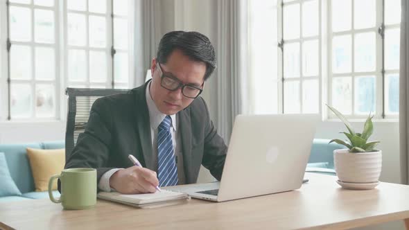 Asian Businessman Typing On Computer Then Taking Note In A Book While Working At Home