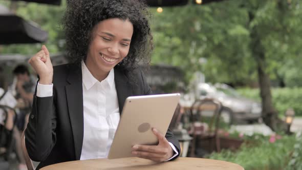 African Woman Celebrating Win on Tablet, Sitting in Outdoor Cafe